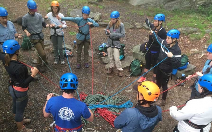 A group of people wearing safety gear stand in a circle and are connected by ropes, likely in an exercise. 